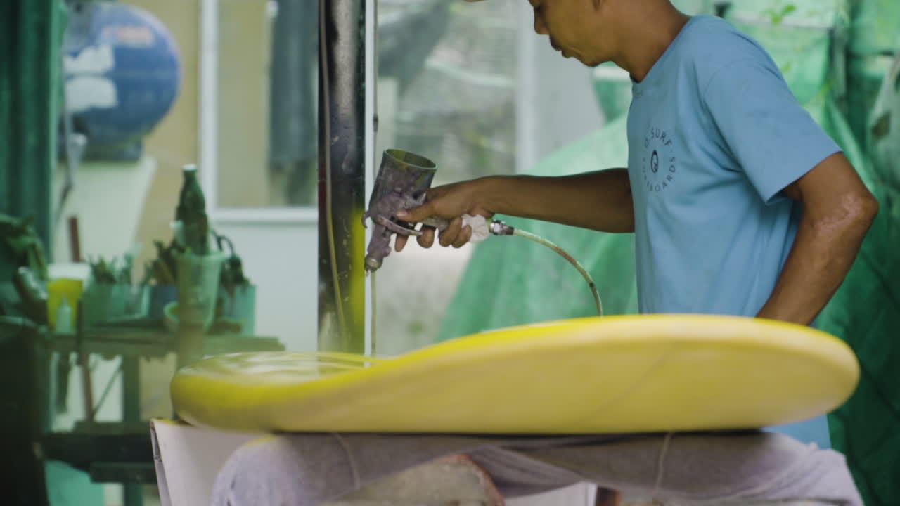 Handyman finishes paint job on surfboard using spray gun with yellow acrylic paint