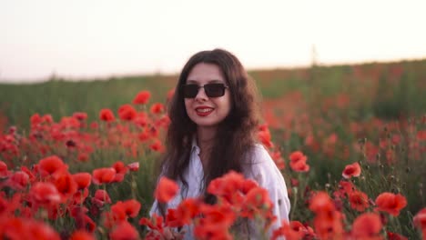 retrato de una hermosa chica de cabello oscuro en un campo de flores silvestres y amapolas rojas, con un vestido y sonriendo mientras se pone gafas de sol