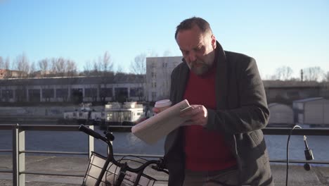 A man with a bicycle leaning against a railing on the embankment reads a newspaper and sips coffee from a cup