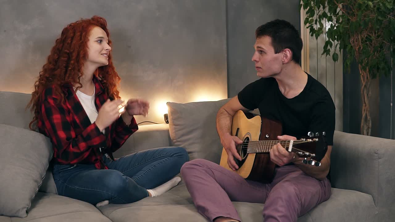 A husband is singing a song and playing a guitar for his wife who is also sitting with him on a sofa and sing with him. She likes to watch him perform. Taking time together at home at living room