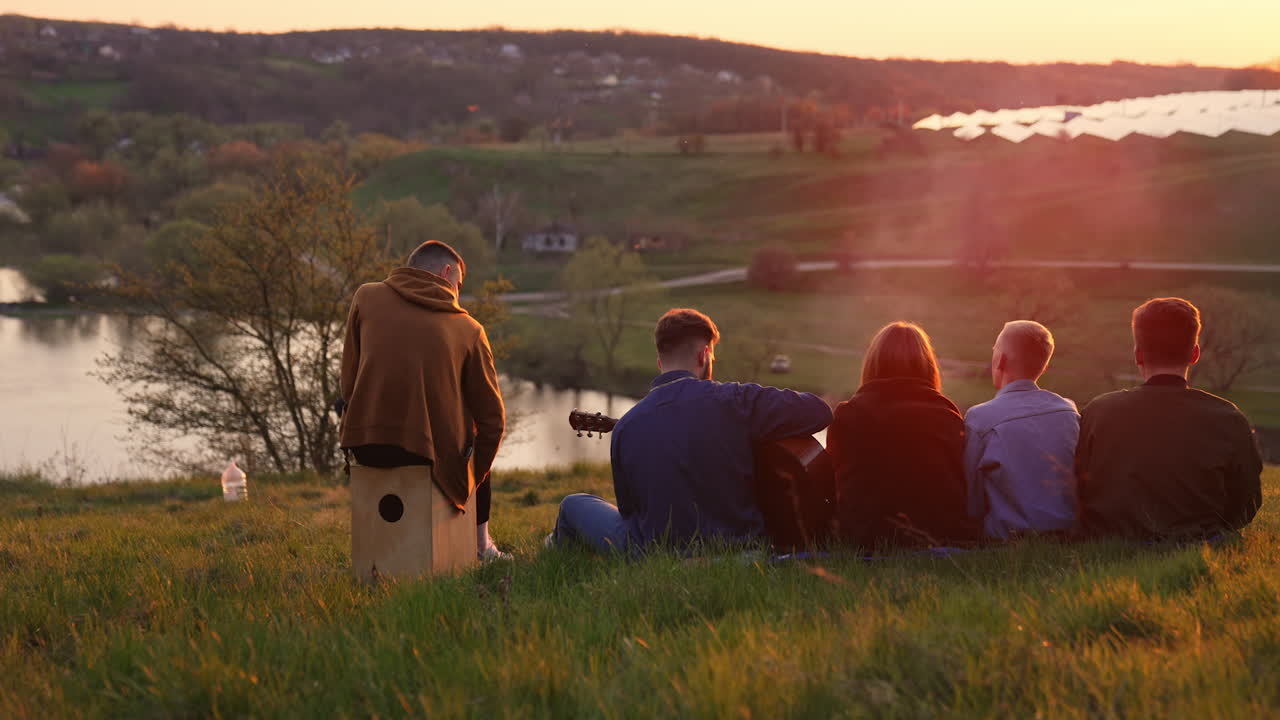 Four young men and woman having good picnic time. Friendly group watching the sunset and singing song along with playing instruments.