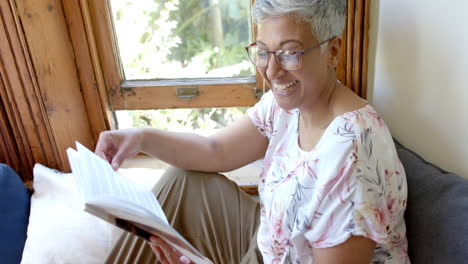 Feliz-Mujer-Birracial-Mayor-En-El-Sofá-Leyendo-Un-Libro-En-La-Ventana-De-Casa,-Cámara-Lenta