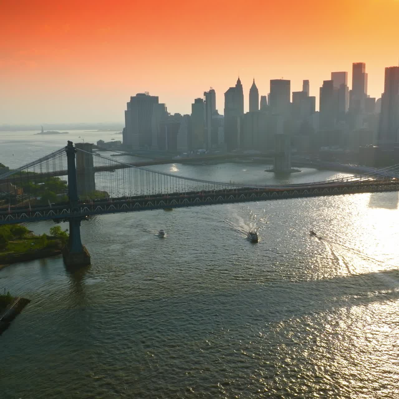 Boats sailing by East River under the beautiful bridges. Silhouettes of famous New York skyscrapers in the rays of setting sun. Pink orange sky backdrop