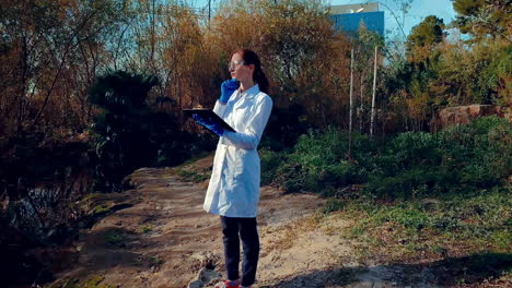 A-young-woman-scientist-at-a-creek,-wearing-protective-eyewear-and-a-lab-coat,-taking-notes