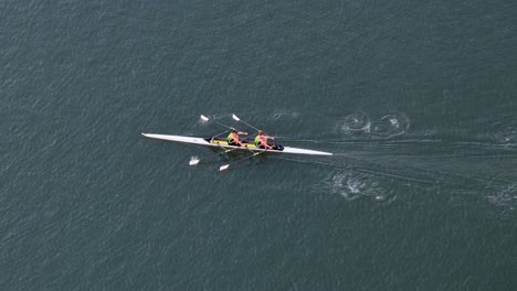 Four-person rowing team practices on calm waters under clear skies, captured from above with smooth camera movement