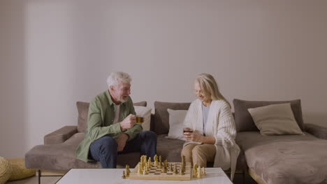 senior man and woman drinking tea and playing chess sitting on sofa at home