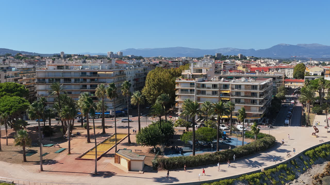 Aerial drone view of the Antibes seafront promenade lined with palm trees and modern apartments, with the Mediterranean Sea and distant mountains in the background