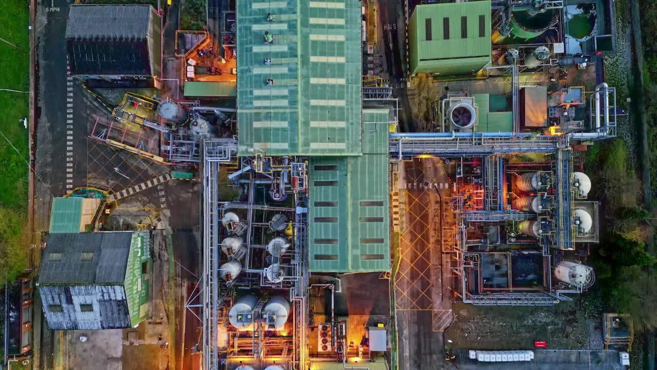 медленная съемка с беспилотника крупного промышленного предприятия в сумерках, на которой видны трубопроводные конструкции, здания, градирни, пар и рабочие машины.