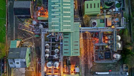 Slow-forward-moving-aerial-dronefootage-of-a-large-industrial-plant-at-dusk,-showing-pipework-structures,-buildings,-cooling-towers,-steam,-and-work-vehicles