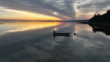 Goldener-Orangefarbener-Sonnenuntergang-über-Dem-Reflektierenden-See-Mit-Einem-Kleinen-Leeren-Boot,-Das-In-Ruhigen-Gewässern-Festgemacht-Ist