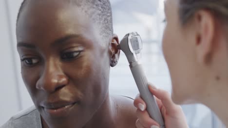 Happy-diverse-female-patient-and-doctor,-checking-her-ear-with-otoscope-in-hospital,-in-slow-motion