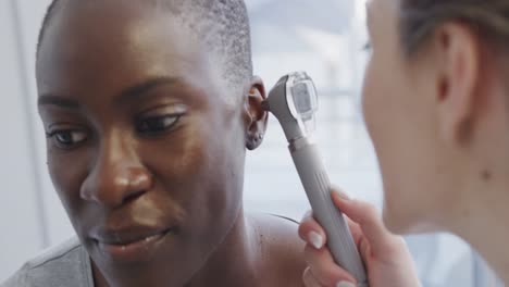 Happy diverse female patient and doctor, checking her ear with otoscope in hospital, in slow motion