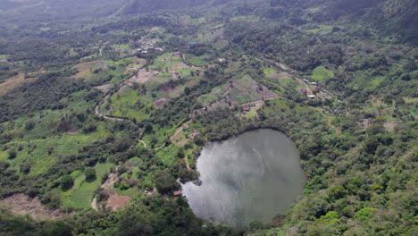 Un-Paisaje-Verde-Y-Exuberante-Con-Un-Lago-Circular,-Rodeado-De-Densos-árboles-Y-Parcelas-De-Tierra-Cultivada,-Vista-Aérea