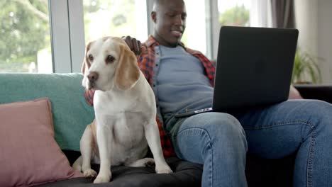 happy african american man sitting on sofa using laptop, with his pet dog at home, slow motion