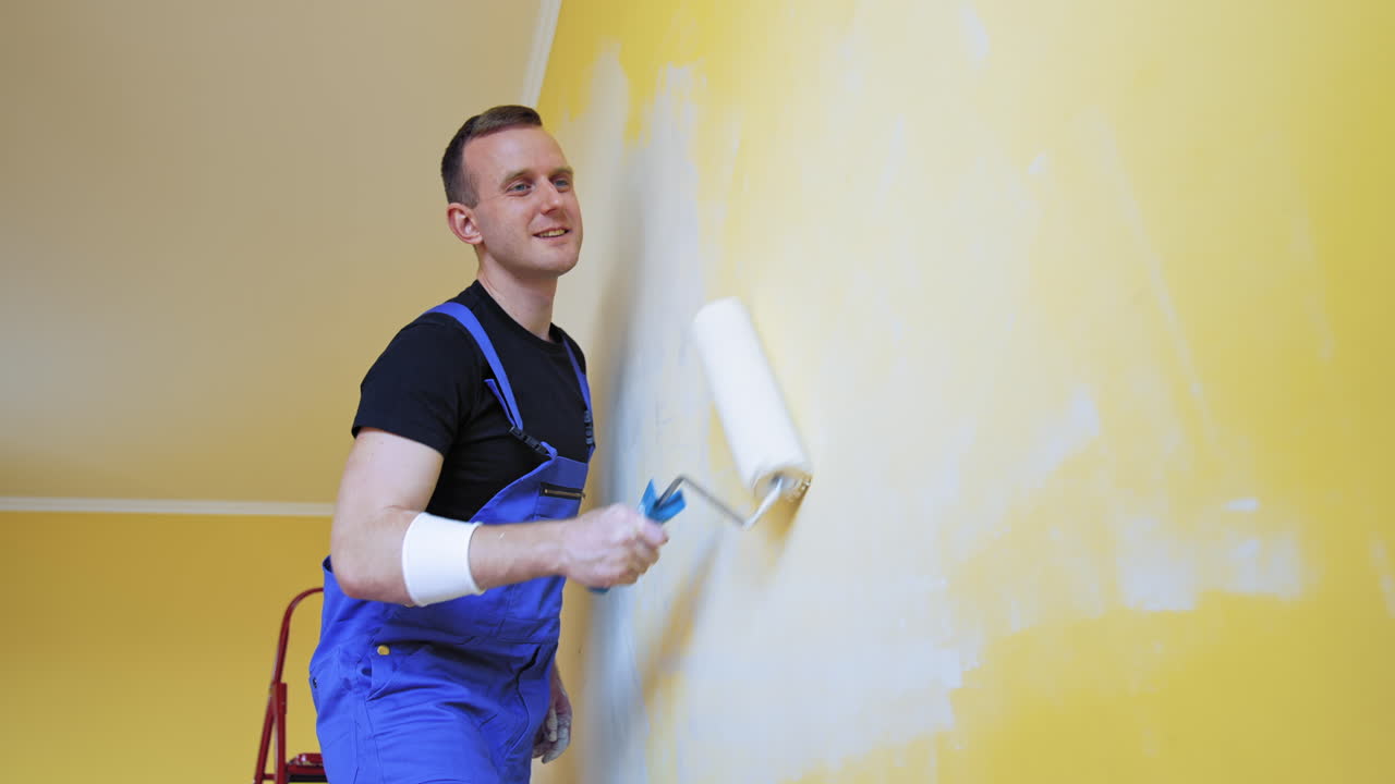Young man improving apartment. Interior decoration in flat. Worker in blue overalls painting wall with a paint roller. Painting services.