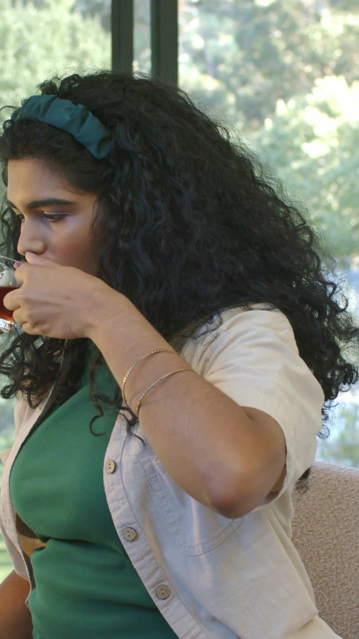 Video verticale: Donna assetata con fascia color verde acqua e braccialetti che solleva una tazza di vetro per sorseggiare vicino alla finestra