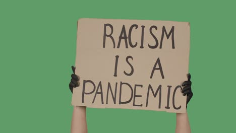 female hands in black gloves pick up a poster from a cardboard box that reads racism is a pandemic. struggle for equality and unity. isolated a green screen, chroma key. close up. slow motion