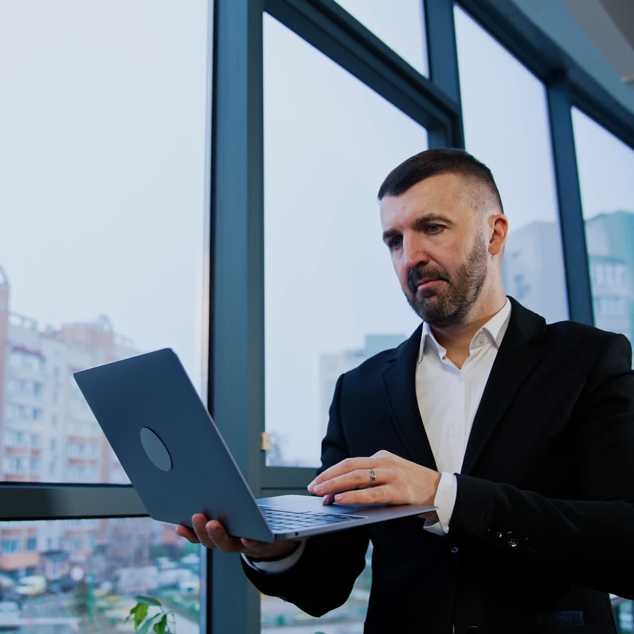 Mid-aged businessman stands at the window holding laptop. Man finishes his work on computer, closes it and looks thoughtfully in the window. Low angle view