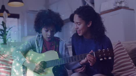 Animation-of-glowing-spots-over-happy-biracial-mother-with-son-playing-guitar