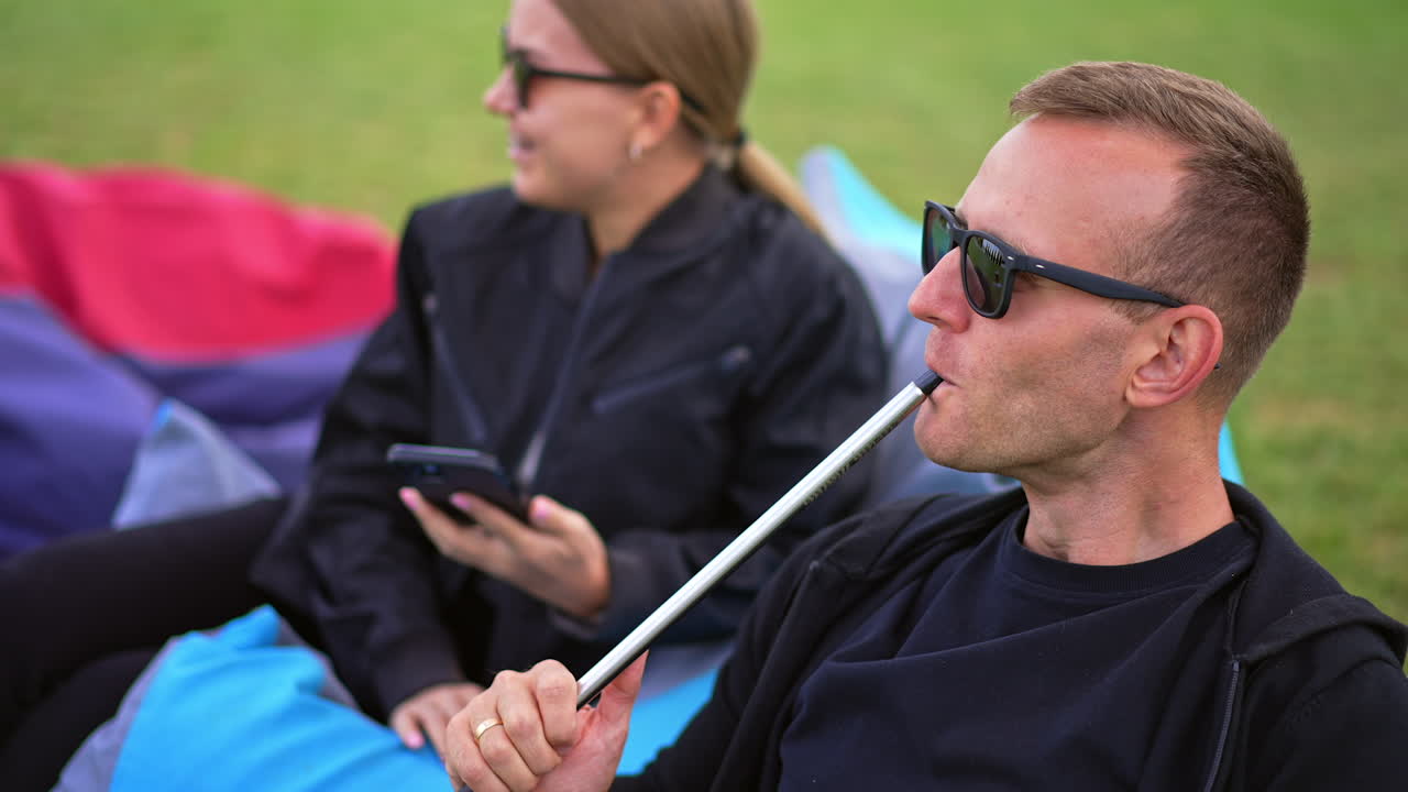 Young couple spending time outdoors resting. Man in sunglasses smoking hookah and woman holding a smartphone and smiling.