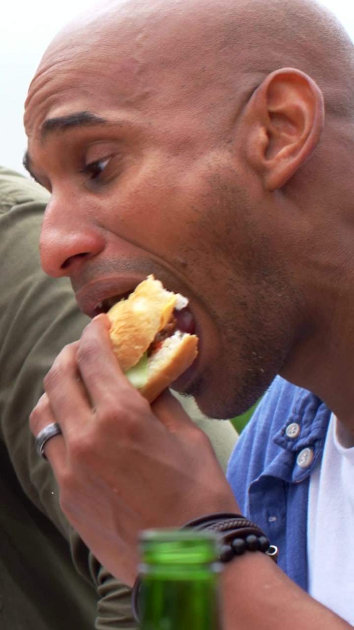 Vertikal video: Snakkende skallet mann i blå skjorte biter i en burger, spiser på en uteplass med en grønn flaske.