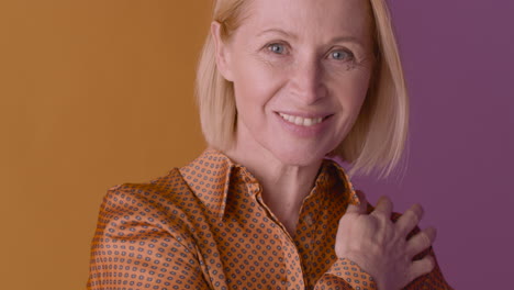 close up view of blonde mature woman wearing brown shirt smiling and touching her shoulder on orange and purple background