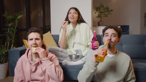 Tres-Amigos-Viendo-Una-Película-Cómica-En-La-Televisión-Sentados-En-El-Sofá-Y-El-Piso,-Comiendo-Papas-Fritas-Y-Palomitas-De-Maíz-Y-Bebiendo-Refrescos