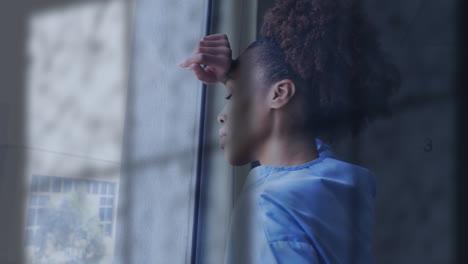 animation of rain on window over unhappy african american female doctor looking out of window