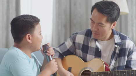 close up of asian father and son with expressive face playing the guitar and singing together on sofa at home