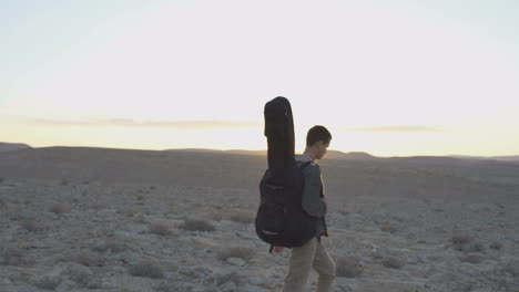 joven camina con estuche de guitarra en el atardecer del desierto