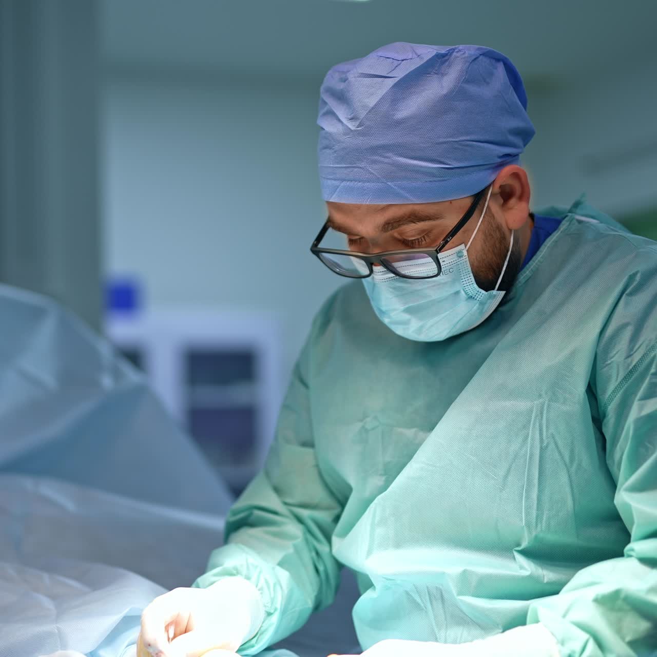 Male plump bearded surgeon wearing glasses does some manipulations with patient's hand. Patient lies covered with sheet. Blurred backdrop