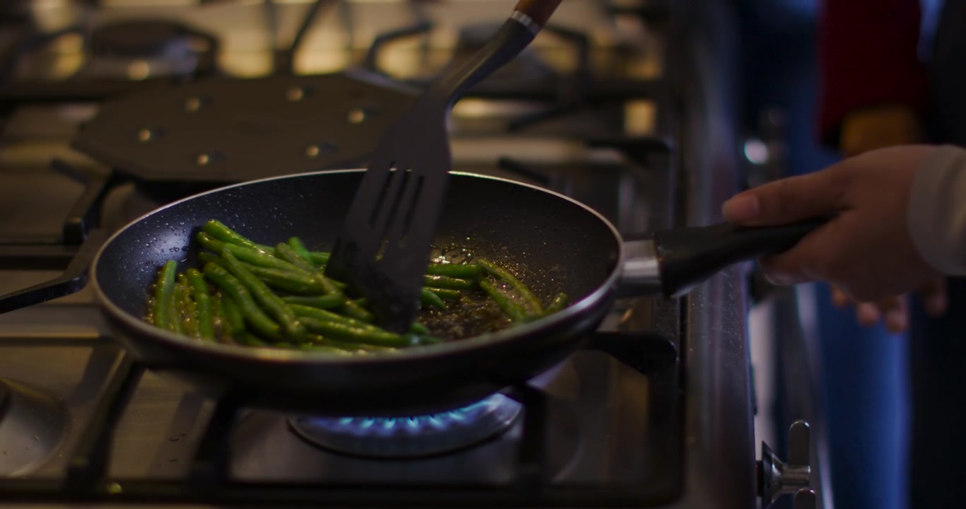 Fiamma a gas che riscalda una padella sul fornello, donna in top rosso che gira fagiolini con una spatola, cucinando uniformemente