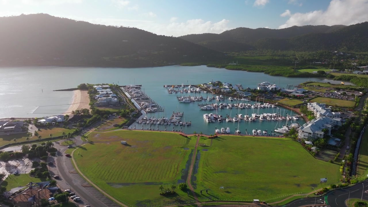 Porto di Airlie Beach, laguna della baia, cuore della Grande Barriera Corallina, Whitsundays, Whitehaven. Ripresa aerea con drone: campo d'erba verde, Mar dei Coralli soleggiato, marina, molo, spiaggia di Boathaven, barche a vela, auto. Movimento in avanti che rivela.