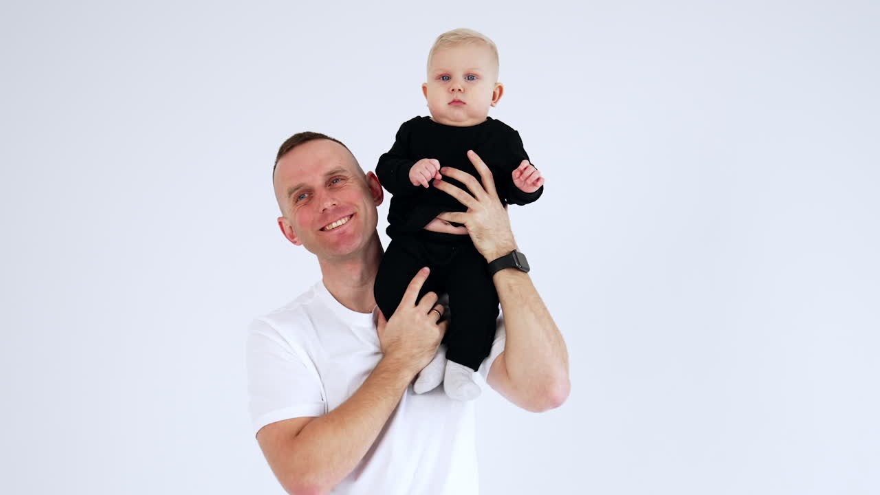 Proud Caucasian man in white t-shirt holding a baby on his shoulder. Calm blond male kid looks peacefully around. White backdrop.