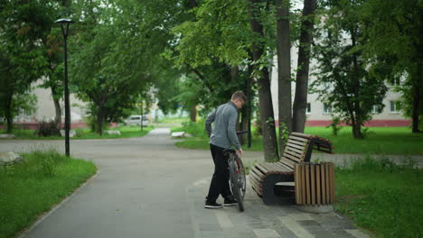 young boy dismounts from bicycle near wooden bench in peaceful, tree-filled park, surrounded by lush greenery, calm environment, sits down, greenery and residential building in background