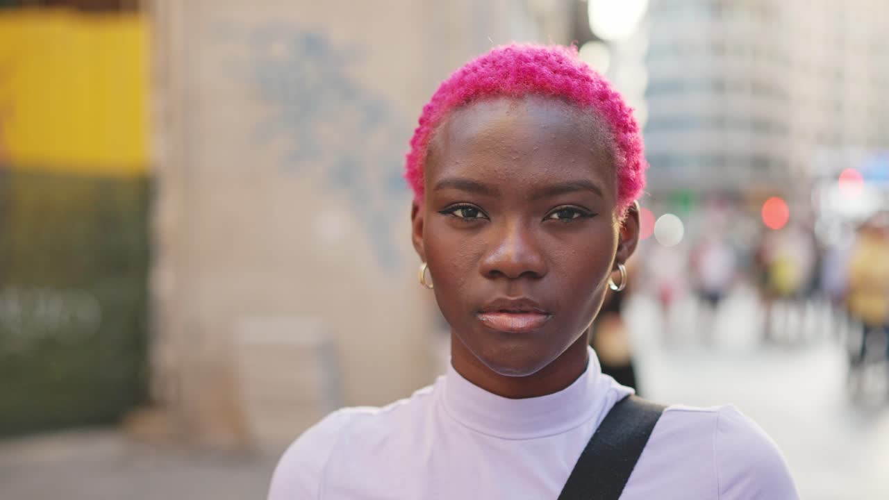 A young woman with pink hair makes a stop gesture with her hand on a city street.