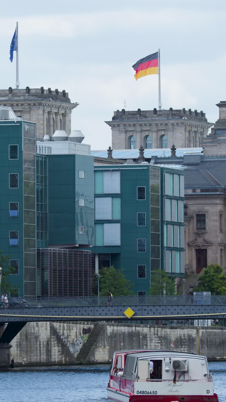 Un barco turístico se desliza por el río, pasando por un paisaje urbano moderno, el Reichstag y banderas alemanas ondeando, bajo una luz diurna nublada.