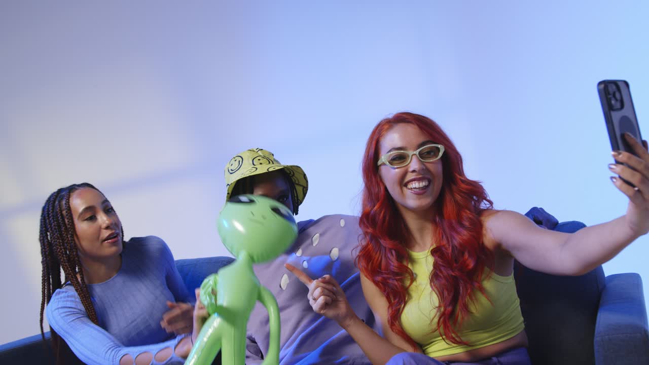 fotografía de estudio de un grupo de amigos de la generación z sentados en el sofá posando para una selfie con un alienígena de juguete en el teléfono móvil 1