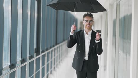 Ein-Glücklicher-Junger-Mann,-Der-Im-Flur-Steht,-öffnet-Einen-Regenschirm-Und-Zeigt-Eine-Plastikkarte