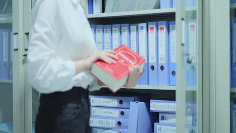 Businesswoman-Looking-For-Required-Document-In-Large-Stack-Of-Papers-On-Bookshelves-In-Office