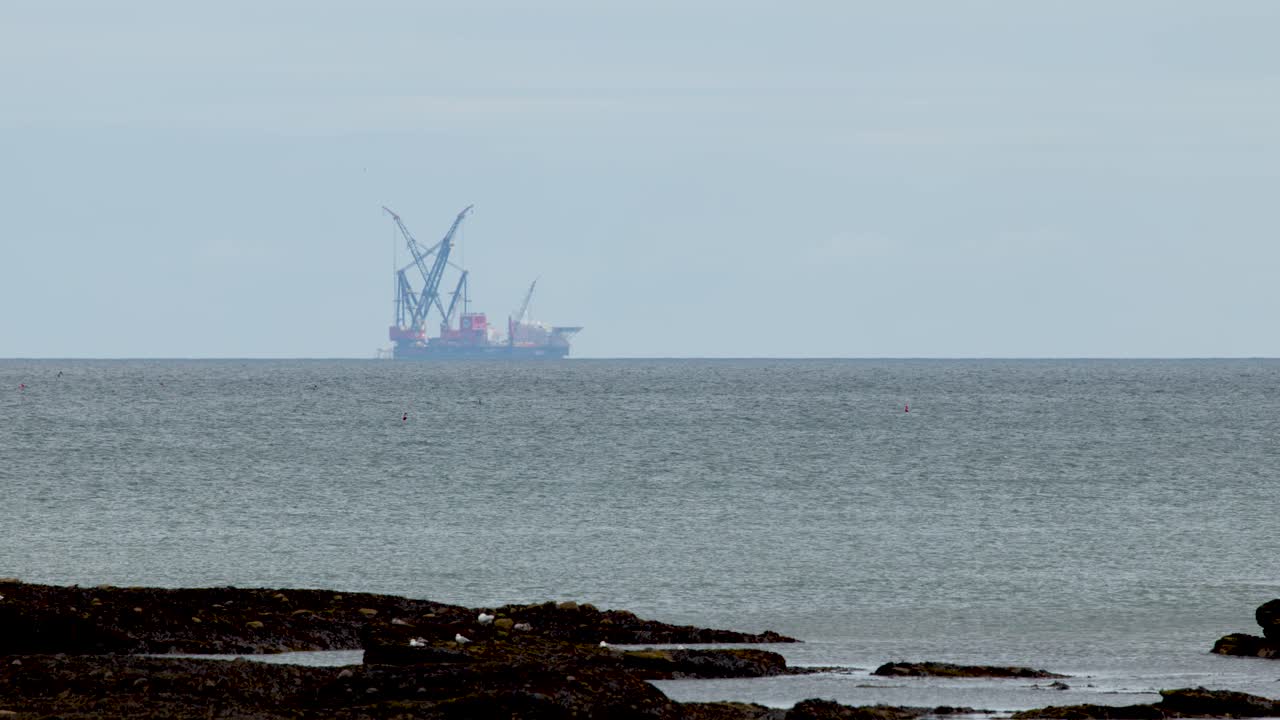 Industrial barge with cranes offshore, calm sea, static wide shot, overcast daylight, Dundee coastline
