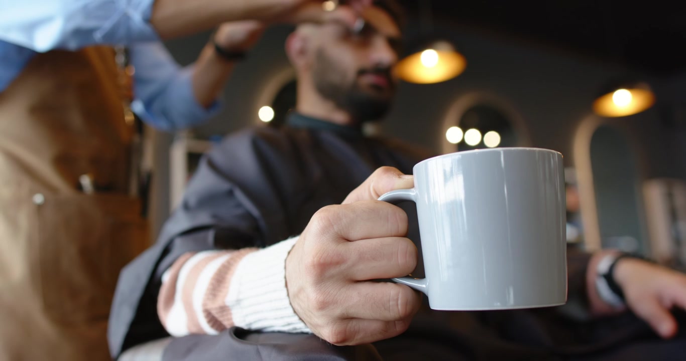 Fotocamera focalizzata su una tazza, con un cliente uomo africano che la tiene in mano e un barbiere uomo che rifinisce i capelli per lo styling.