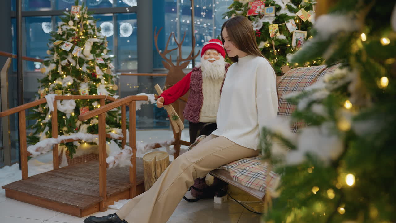 Young woman walking up to a decorative seat with Christmas tree and Santa Claus statue in background, surrounded by festive holiday decorations and warm lights