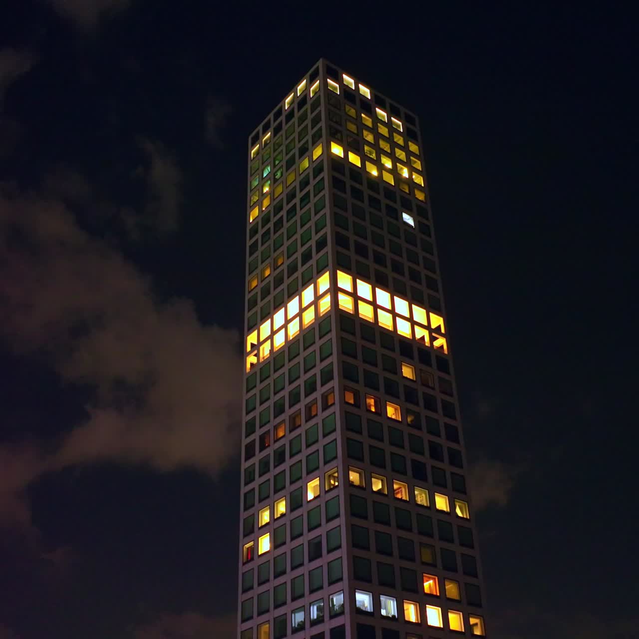 Top of the skyscraper with lights in the windows. Drone footage rising along the building at the backdrop of dark night sky with clouds