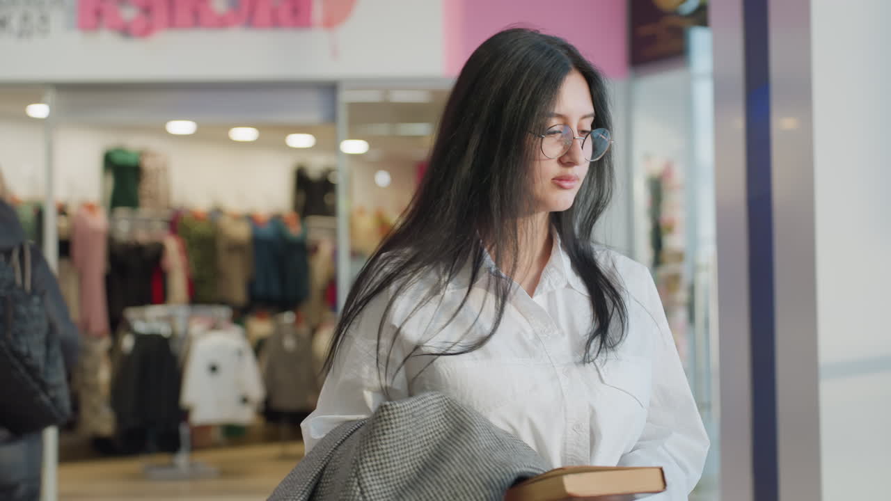 Lady holding book and jacket walks inside modern shopping mall toward decorative bench near indoor plants, drops jacket gently, sits down, and begins reading book with clothing store in background
