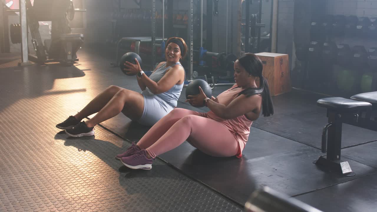 Duo femminile eterogeneo che indossa abbigliamento da allenamento, reclinandosi e torcendo il busto su un tappetino in palestra con una palla medica, tonificando gli addominali.