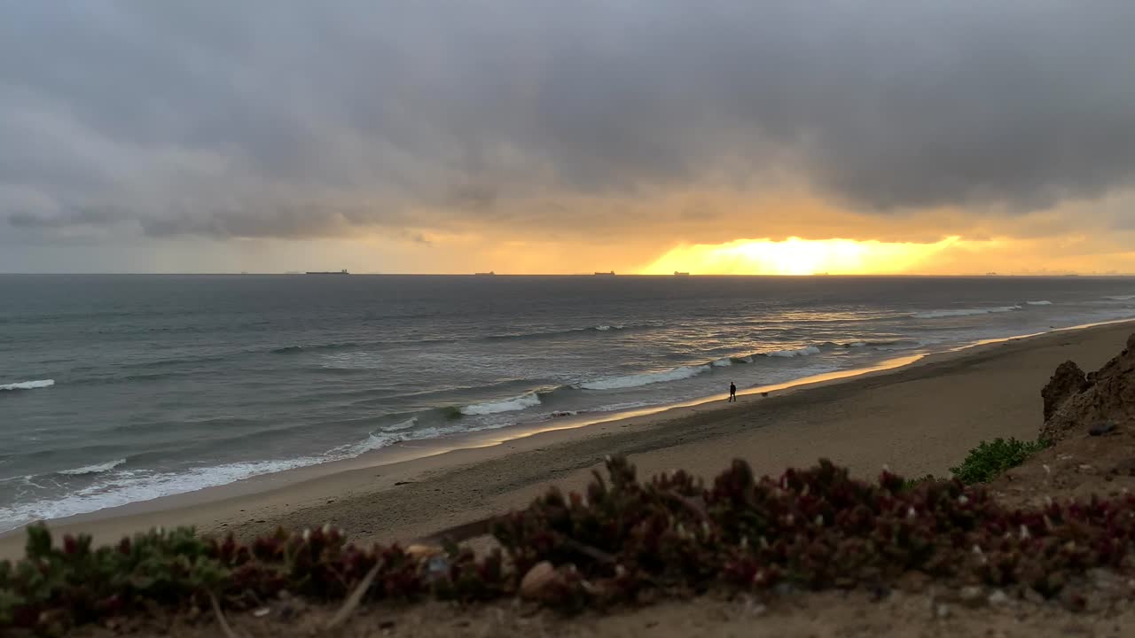 4k 60p, puesta de sol dorada sobre las olas del océano lamiendo en una playa con un hombre paseando a su perro