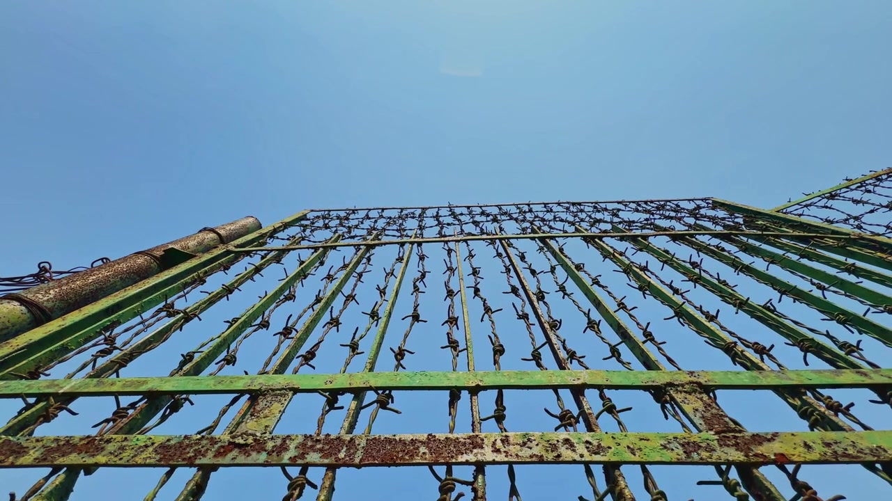 Vista cinematográfica em ângulo baixo e em movimento de um portão alto de metal verde reforçado com fios de arame farpado enferrujado estendendo-se para cima contra um céu azul brilhante em uma área externa segura.