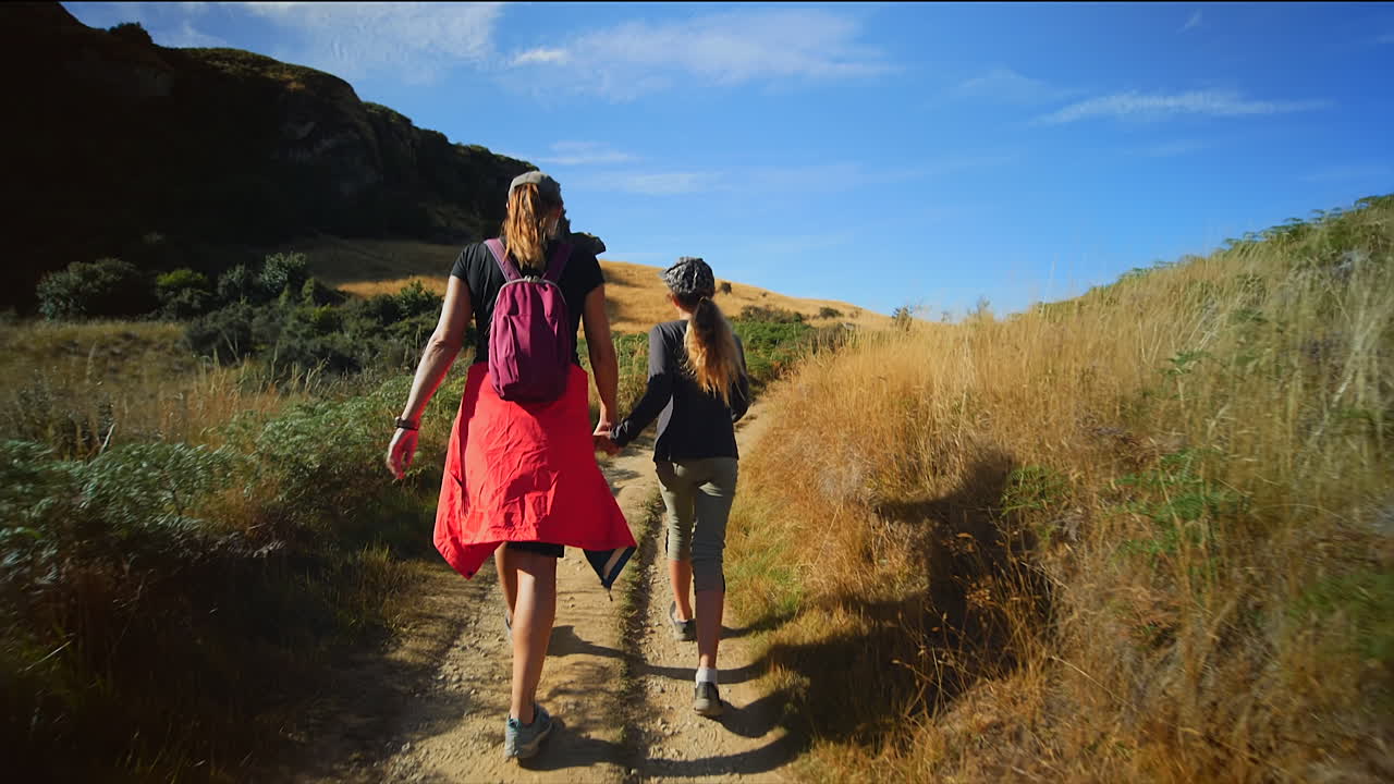 Mother and daughter hike on path in hilly New Zealand landscape, slomo from behind
