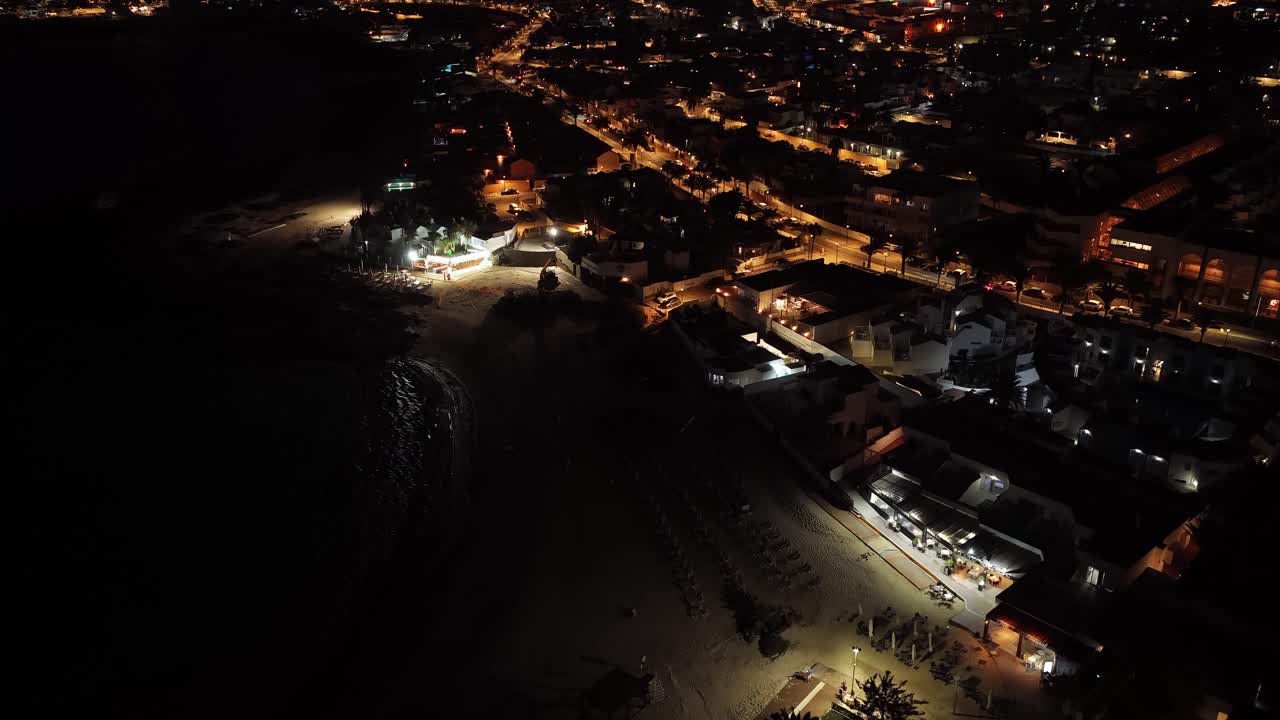Drone glides over Corralejo Viejo at night, revealing a glowing coastal town with beachfront symmetry, streetlights outlining urban geometry, and the dark Atlantic forming a dramatic visual contrast